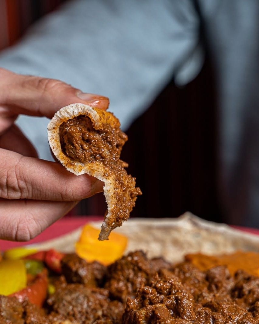 Una mano solleva un boccone di injera arrotolata sopra un vassoio di stufato.